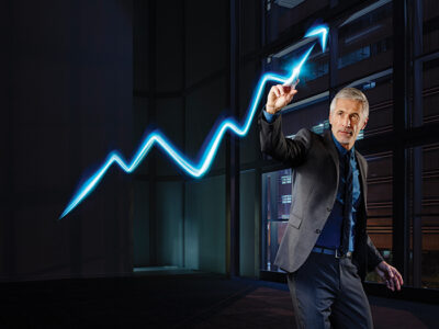 Image of a professional older man in a suit draws a glowing upward-trending line in the air, symbolizing growth and success, with a modern office building in the background.
