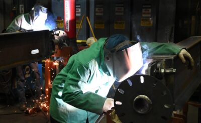 A graphic image of two workers wearing protective welding helmets and gloves as they weld large metal components in an industrial workshop, with bright sparks and smoke visible around the equipment.