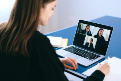 A woman sits at a desk participating in a virtual meeting on a laptop. On the screen, three other participants appear in separate video windows. She holds a pen and looks down at a sheet of paper, as if taking notes. A notebook rests beside the laptop on a blue desk, and the setting appears to be a quiet workspace.